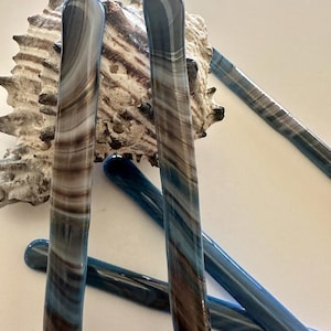 May include: Several glass stir sticks in shades of blue, brown, and white are arranged against a seashell. The glass rods have a rounded top and a swirled pattern. The seashell is beige and has a textured surface.