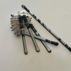 May include: A collection of black and white hair pins, some straight and some curved, are arranged near a textured seashell. The pins have a striped pattern and rounded tips. One long pin is also visible.