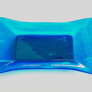 Op de afbeelding: Een rechthoekige, blauwe glazen schaal met een gebogen, vlinderdasvorm. De schaal heeft een donkerblauw rechthoekig midden en lichtblauwe randen, waardoor een gradiënteffect ontstaat. Het glas lijkt doorschijnend.