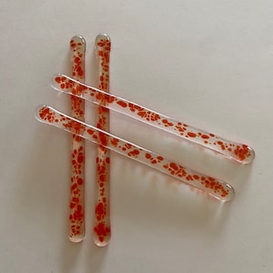 May include: Four clear glass stir sticks with rounded ends. Each stick is decorated with orange speckled patterns. The sticks are arranged diagonally against a white background, showcasing their design and clarity.