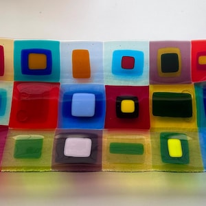 May include: A rectangular fused glass tray featuring a grid of colorful squares and rectangles. The design includes translucent and opaque glass segments in shades of red, blue, yellow, green, orange, purple, and white, arranged in a mosaic pattern.