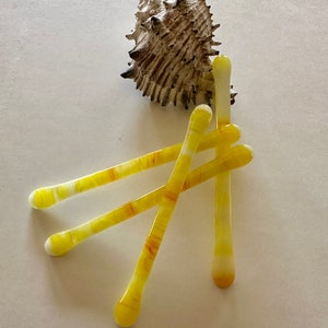 Può includere: Una natura morta con una conchiglia beige strutturata e diversi fermagli per capelli in vetro giallo con striature bianche e arancioni. I fermagli sono disposti a grappolo, con punte arrotondate.