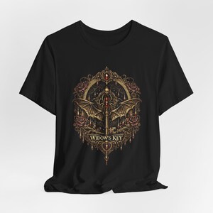 Op de afbeelding: Zwart T-shirt met een gedetailleerd grafisch ontwerp. Het ontwerp bevat een sleutel met vleermuisvleugels, een schedel, rozen en de tekst "Widow's Key" in een decoratief kader. Het kunstwerk gebruikt goud-, rood- en bruintinten.