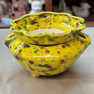 Może przedstawiać: Jaskrawożółta ceramiczna doniczka z falistym brzegiem i nakrapianym wzorem w kolorze niebieskim i fioletowym. Naczynie w kształcie misy posiada trzy małe nóżki u podstawy oraz dwa boczne uchwyty, z gładkim, błyszczącym wykończeniem na zewnątrz.