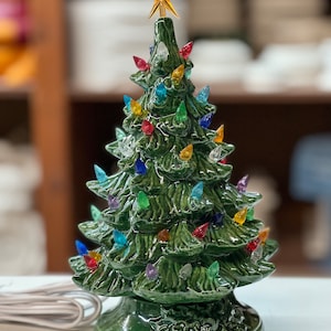 以下が含まれることがあります： 光沢のある緑色のセラミッククリスマスツリー。カラフルなライトと金の星で飾られています。ベースには装飾的なデザインがあります。白い電気ケーブルが取り付けられています。