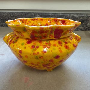 Op de afbeelding: Een tweedelige keramische bowls set. De bowls zijn geel met een gespikkeld patroon van rood, oranje en groen. De bovenste bowl is kleiner en heeft een golvende rand. De onderste bowl is groter en heeft drie kleine voetjes.