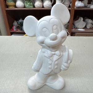 以下が含まれることがあります： 白いセラミック製のミッキーマウスの置物。スーツを着て帽子を持っています。大きな耳、丸い鼻、笑顔の表情をしています。未塗装で、カスタマイズできます。