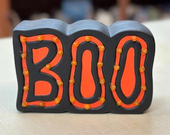 以下が含まれることがあります： オレンジ色の「BOO」の文字が小さなオレンジ色のライトで縁取られた黒いハロウィーンの装飾。文字はオレンジ色の背景にあります。装飾は明るい色の表面にあります。
