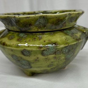 May include: A handcrafted ceramic container with a lid. The container is a vibrant green with darker green and brown speckled accents. The lid sits atop the bowl-shaped base. The container has three small feet.