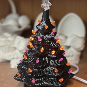 Op de afbeelding: Een zwarte keramische Halloween-boom met oranje en roze lampjes. De boom is bekroond met een doorzichtig spookvormig ornament. De boom is aangesloten met een wit snoer. Een feestelijke decoratie voor het seizoen.