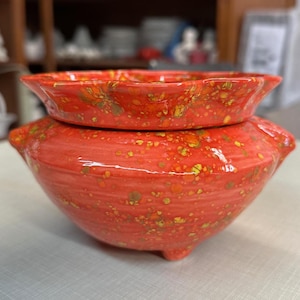 May include: A two-piece ceramic pottery set in a vibrant red hue with speckled yellow and green accents. The top piece is a shallow dish, and the bottom is a rounded bowl with three small feet. The set has a glossy finish.