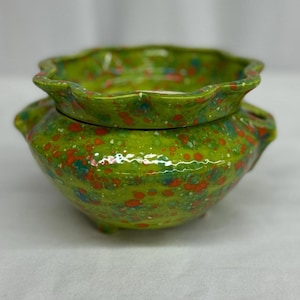 Op de afbeelding: Een keramische pot met deksel, in een levendige groene kleur, bespikkeld met rode, blauwe en witte stippen. De rand heeft een geschulpte rand. De pot heeft kleine handvatten en staat op drie kleine voetjes.