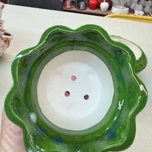 Könnte beinhalten: Ein grüner, blumenförmiger Keramik-Pflanztopf mit weißem Innenraum. Der Pflanztopf hat eine glänzende Oberfläche mit blauen und weißen Sprenkeln. Am Boden des Pflanztopfs sind drei Abzugslöcher zu sehen.