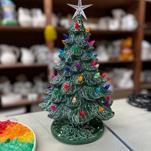 Op de afbeelding: Een keramische kerstboom, ongeveer 30 cm hoog, met een groene textuurafwerking, versierd met kleurrijke lichtjes en een ster als piek. De boom staat op een groene basis met een hulstbladontwerp. Een feestelijke vakantiedecoratie.