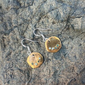 May include: A pair of gold-toned earrings with small green and blue stones set in the round metal discs. The earrings have silver hooks.