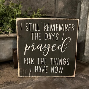 May include: A square, black wooden sign with white lettering. The text reads, "I STILL REMEMBER THE DAYS I prayed FOR THE THINGS I HAVE NOW." The sign has a distressed, rustic look with a white border.
