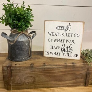 May include: A rustic wooden box holds a galvanized metal vase with greenery and a small wooden sign. The sign reads "accept what is, let go of what was, have faith in what will be." The vase is tied with twine.