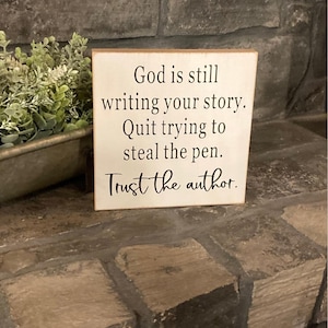 May include: A small, square, off-white wooden sign with black text. The text reads: "God is still writing your story. Quit trying to steal the pen. Trust the author." The sign is placed on a stone surface.