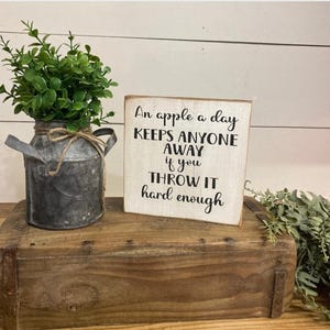 May include: A rustic wooden box holds a small metal milk jug with greenery and a wooden sign. The sign reads "An apple a day keeps anyone away if you throw it hard enough" in black text on a white background.