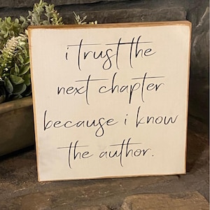 May include: A square wooden sign with a cream-colored background and a brown border. The sign features black cursive text that reads, "I trust the next chapter because I know the author."