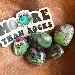 May include: A collection of polished zoisite stones with green and pink hues, displayed in a wooden bowl. A sticker with the text "MORE THAN ROCKS" and a globe graphic is also present.