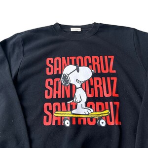 May include: Black sweatshirt with a red "Santa Cruz" logo repeated three times. A cartoon image of Snoopy wearing sunglasses and riding a skateboard is in the center of the sweatshirt.