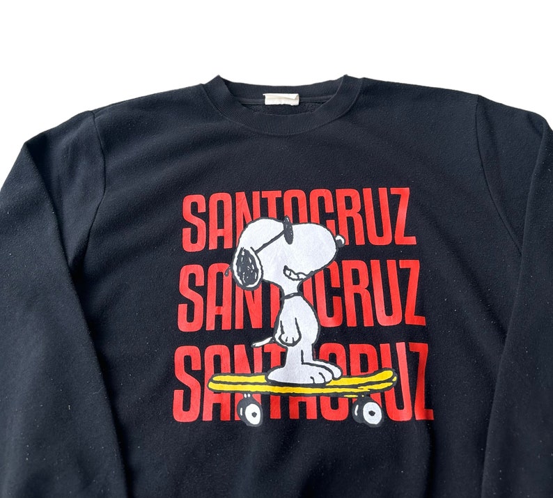 May include: Black sweatshirt with a red "Santa Cruz" logo repeated three times. A cartoon image of Snoopy wearing sunglasses and riding a skateboard is in the center of the sweatshirt.