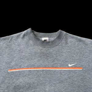 K&ouml;nnte beinhalten: Ein graues Sweatshirt mit einem wei&szlig;en Nike-Swoosh und einem orangefarbenen Streifen &uuml;ber der Brust. Das Nike-Logo ist am Kragen sichtbar.