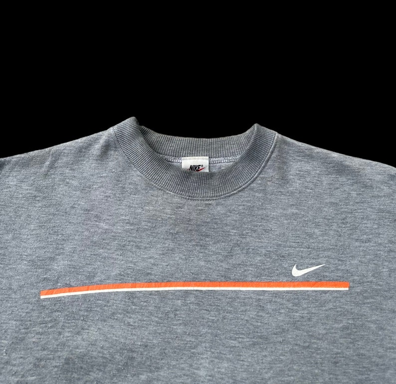 K&ouml;nnte beinhalten: Ein graues Sweatshirt mit einem wei&szlig;en Nike-Swoosh und einem orangefarbenen Streifen &uuml;ber der Brust. Das Nike-Logo ist am Kragen sichtbar.
