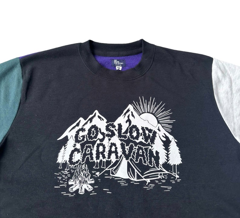 Puede incluir: Sudadera negra con un gr&aacute;fico blanco de una cordillera y el texto "Go Slow Caravan". La sudadera tiene paneles de color verde y gris en las mangas.