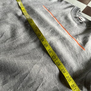 K&ouml;nnte beinhalten: Graues T-Shirt mit einem d&uuml;nnen orangenen Streifen &uuml;ber der Brust. Das Shirt wird mit einem gelben Ma&szlig;band gemessen.