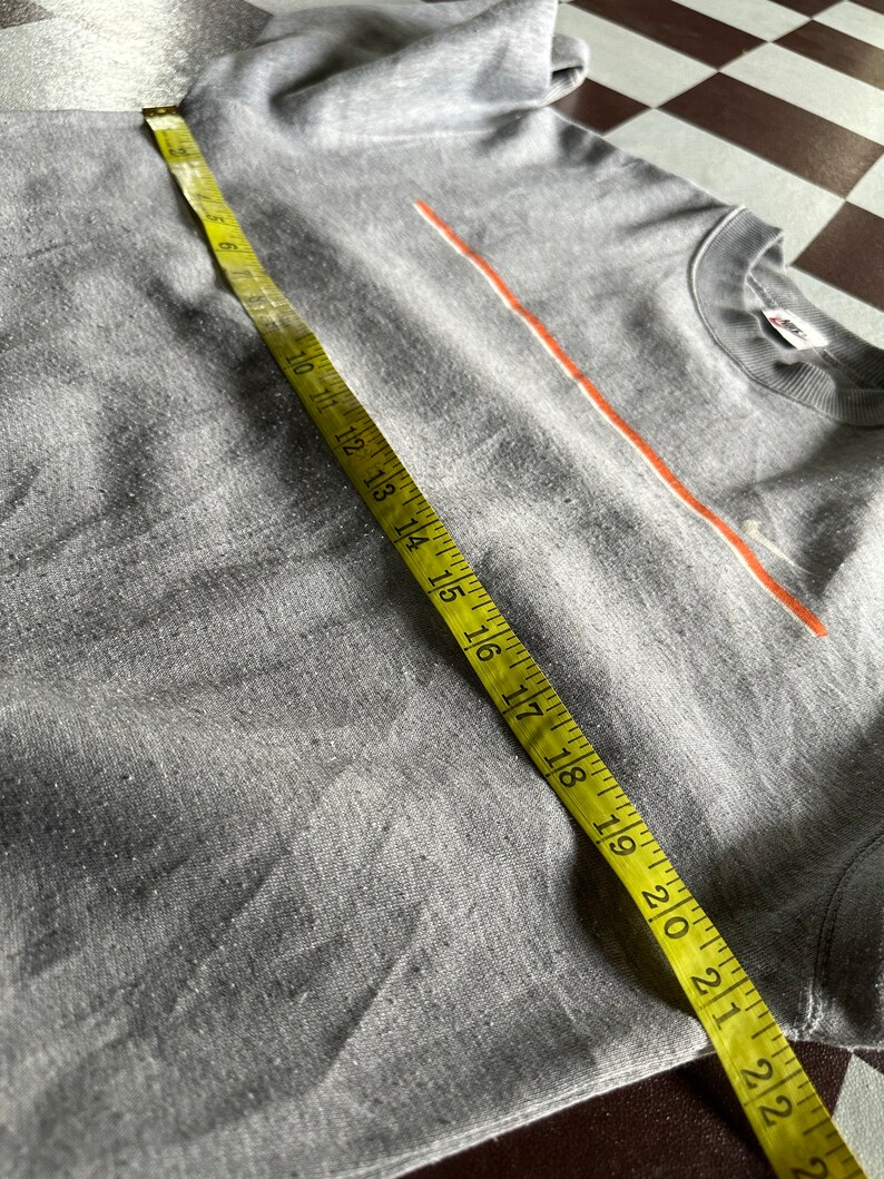 K&ouml;nnte beinhalten: Graues T-Shirt mit einem d&uuml;nnen orangenen Streifen &uuml;ber der Brust. Das Shirt wird mit einem gelben Ma&szlig;band gemessen.