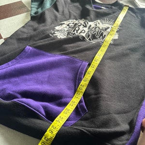 Puede incluir: Sudadera negra y morada con un estampado gr&aacute;fico blanco de una cordillera y la palabra "Slatn" en la parte delantera. La sudadera tiene un bolsillo en la parte delantera y una cinta m&eacute;trica est&aacute; colocada sobre la parte delantera de la sudadera.