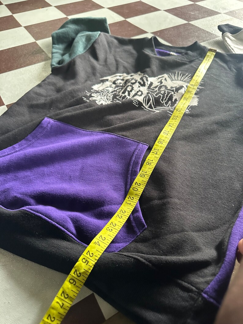 Puede incluir: Sudadera negra y morada con un estampado gr&aacute;fico blanco de una cordillera y la palabra "Slatn" en la parte delantera. La sudadera tiene un bolsillo en la parte delantera y una cinta m&eacute;trica est&aacute; colocada sobre la parte delantera de la sudadera.