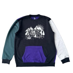 Puede incluir: Una sudadera negra, verde y gris con un bolsillo morado. La sudadera tiene un estampado gr&aacute;fico de una escena de monta&ntilde;a con el texto "Go Slow Caravan".