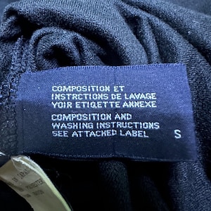 K&ouml;nnte beinhalten: Nahaufnahme eines dunkelblauen Kleidungsst&uuml;cketiketts mit wei&szlig;em Text. Das Etikett enth&auml;lt die W&ouml;rter "COMPOSITION ET INSTRCTIONS DE LAVAGE VOIR ETIGETTE ANNEXE" und "COMPOSITION AND WASHING INSTRUCTIONS SEE ATTACHED LABEL". Der Buchstabe "S" ist sichtbar.
