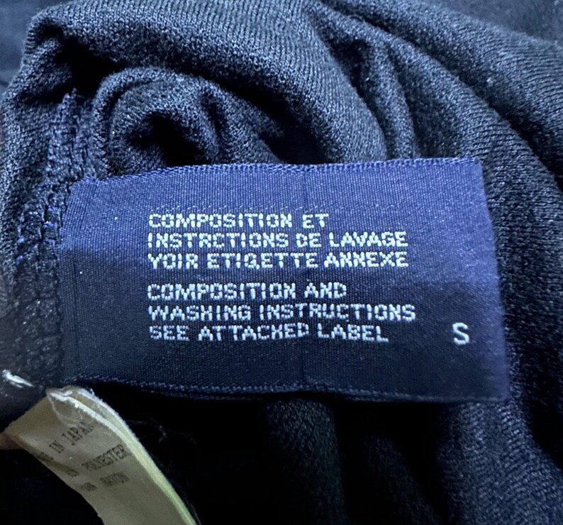 K&ouml;nnte beinhalten: Nahaufnahme eines dunkelblauen Kleidungsst&uuml;cketiketts mit wei&szlig;em Text. Das Etikett enth&auml;lt die W&ouml;rter "COMPOSITION ET INSTRCTIONS DE LAVAGE VOIR ETIGETTE ANNEXE" und "COMPOSITION AND WASHING INSTRUCTIONS SEE ATTACHED LABEL". Der Buchstabe "S" ist sichtbar.