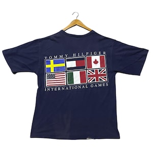 May include: A navy blue Tommy Hilfiger t-shirt displaying flags of Sweden, Germany, Canada, the United States, Italy, and the United Kingdom. The text "TOMMY HILFIGER INTERNATIONAL GAMES" is printed above and below the flags. The shirt is hanging on a wooden hanger.