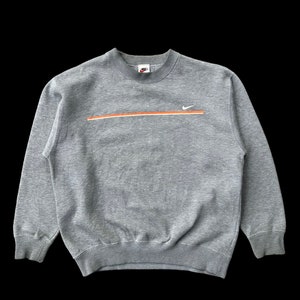 K&ouml;nnte beinhalten: Ein grauer Pullover mit einem wei&szlig;en Nike-Swoosh und einem orangefarbenen Streifen &uuml;ber der Brust. Der Pullover hat einen Rundhalsausschnitt und lange &Auml;rmel.
