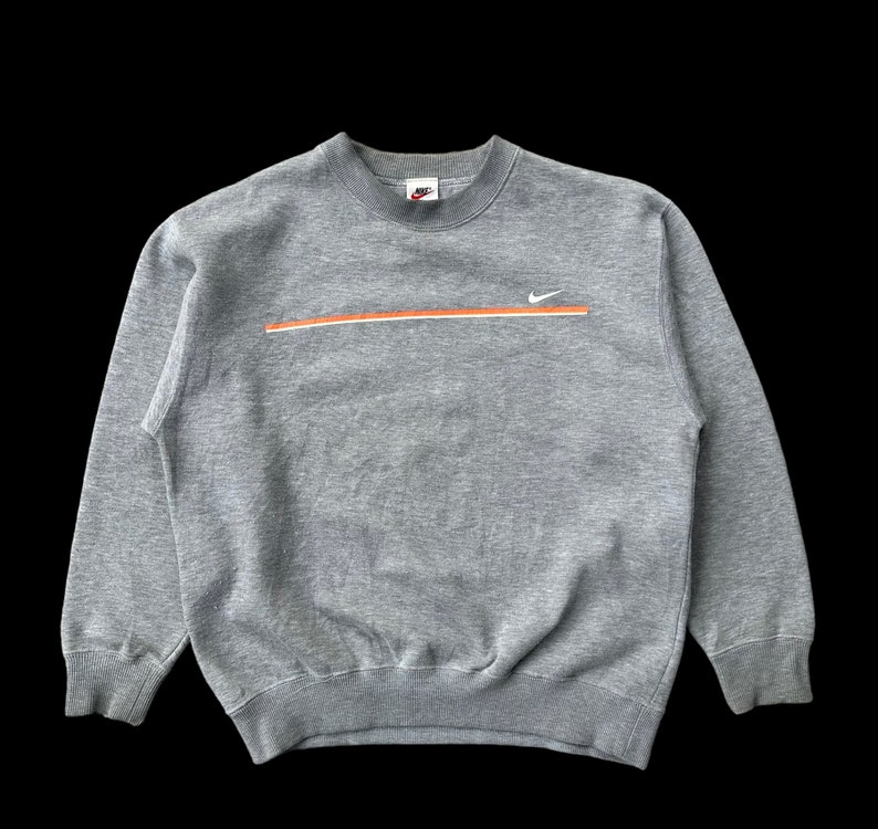 K&ouml;nnte beinhalten: Ein grauer Pullover mit einem wei&szlig;en Nike-Swoosh und einem orangefarbenen Streifen &uuml;ber der Brust. Der Pullover hat einen Rundhalsausschnitt und lange &Auml;rmel.
