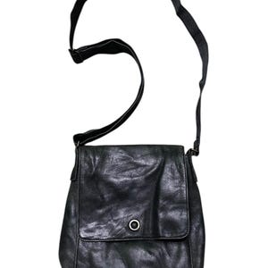 May include: Black leather crossbody bag with a flap closure and adjustable strap. The bag features a circular metal detail on the front. The leather appears worn, giving it a vintage look. The strap is attached to the bag with metal hardware.