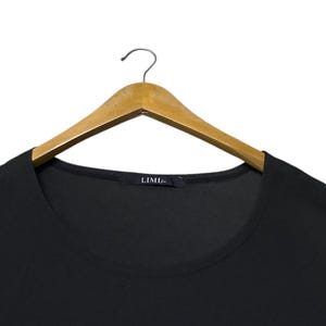 K&ouml;nnte beinhalten: Ein schwarzes Oberteil mit Rundhalsausschnitt, das an einem Holzkleiderb&uuml;gel h&auml;ngt. Das Oberteil hat ein schlichtes, minimalistisches Design. Der Markenname "LIMI" ist auf einem kleinen Etikett am Ausschnitt zu sehen.