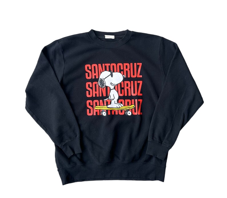 May include: Black sweatshirt with a red "Santa Cruz" logo and a cartoon image of Snoopy wearing sunglasses and riding a skateboard.