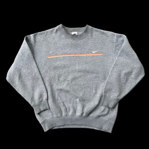 K&ouml;nnte beinhalten: Ein grauer Pullover mit einem wei&szlig;en Nike-Swoosh und einem orangefarbenen Streifen &uuml;ber der Brust.