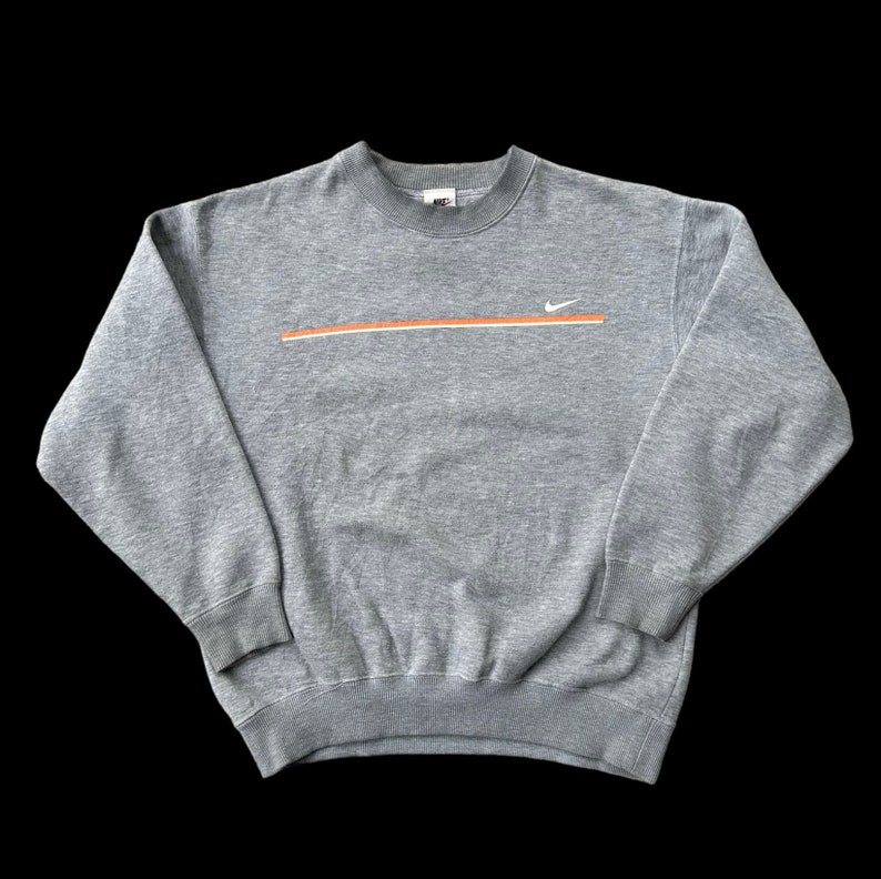 K&ouml;nnte beinhalten: Ein grauer Pullover mit einem wei&szlig;en Nike-Swoosh und einem orangefarbenen Streifen &uuml;ber der Brust.