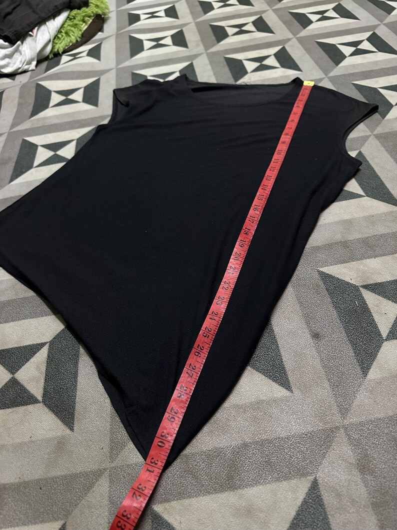 K&ouml;nnte beinhalten: Ein schwarzes, &auml;rmelloses Oberteil mit asymmetrischem Saum. Ein rotes Ma&szlig;band zeigt eine L&auml;nge von etwa 84 cm. Das Oberteil hat einen Rundhalsausschnitt und ein schlichtes, minimalistisches Design.