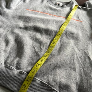 K&ouml;nnte beinhalten: Grauer Sweatshirt mit einem d&uuml;nnen orangenen Streifen &uuml;ber der Brust. Der Sweatshirt hat einen gerippten Saum und B&uuml;ndchen. Ein gelbes Ma&szlig;band ist um den unteren Teil des Sweatshirts gewickelt.