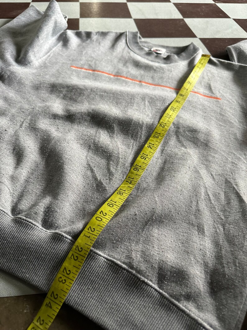 K&ouml;nnte beinhalten: Grauer Sweatshirt mit einem d&uuml;nnen orangenen Streifen &uuml;ber der Brust. Der Sweatshirt hat einen gerippten Saum und B&uuml;ndchen. Ein gelbes Ma&szlig;band ist um den unteren Teil des Sweatshirts gewickelt.