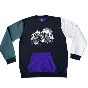 Puede incluir: Una sudadera negra, verde y gris con un bolsillo morado. La sudadera tiene un gr&aacute;fico de monta&ntilde;as y el texto "Go Slow Caravan".