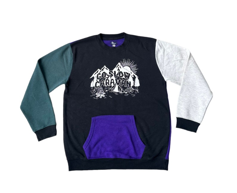 Puede incluir: Una sudadera negra, verde y gris con un bolsillo morado. La sudadera tiene un gr&aacute;fico de monta&ntilde;as y el texto "Go Slow Caravan".